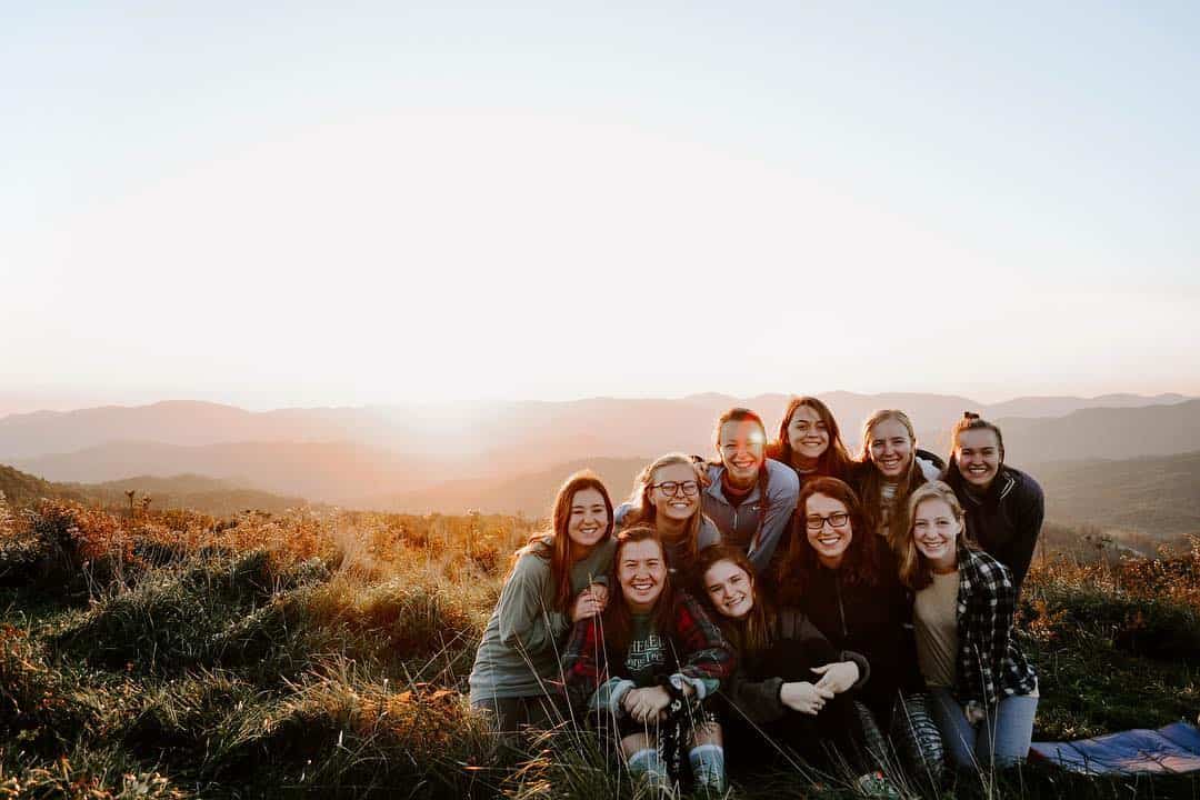 A group of girls gathered, embracing each other in front of a setting sun in an outdoor scene