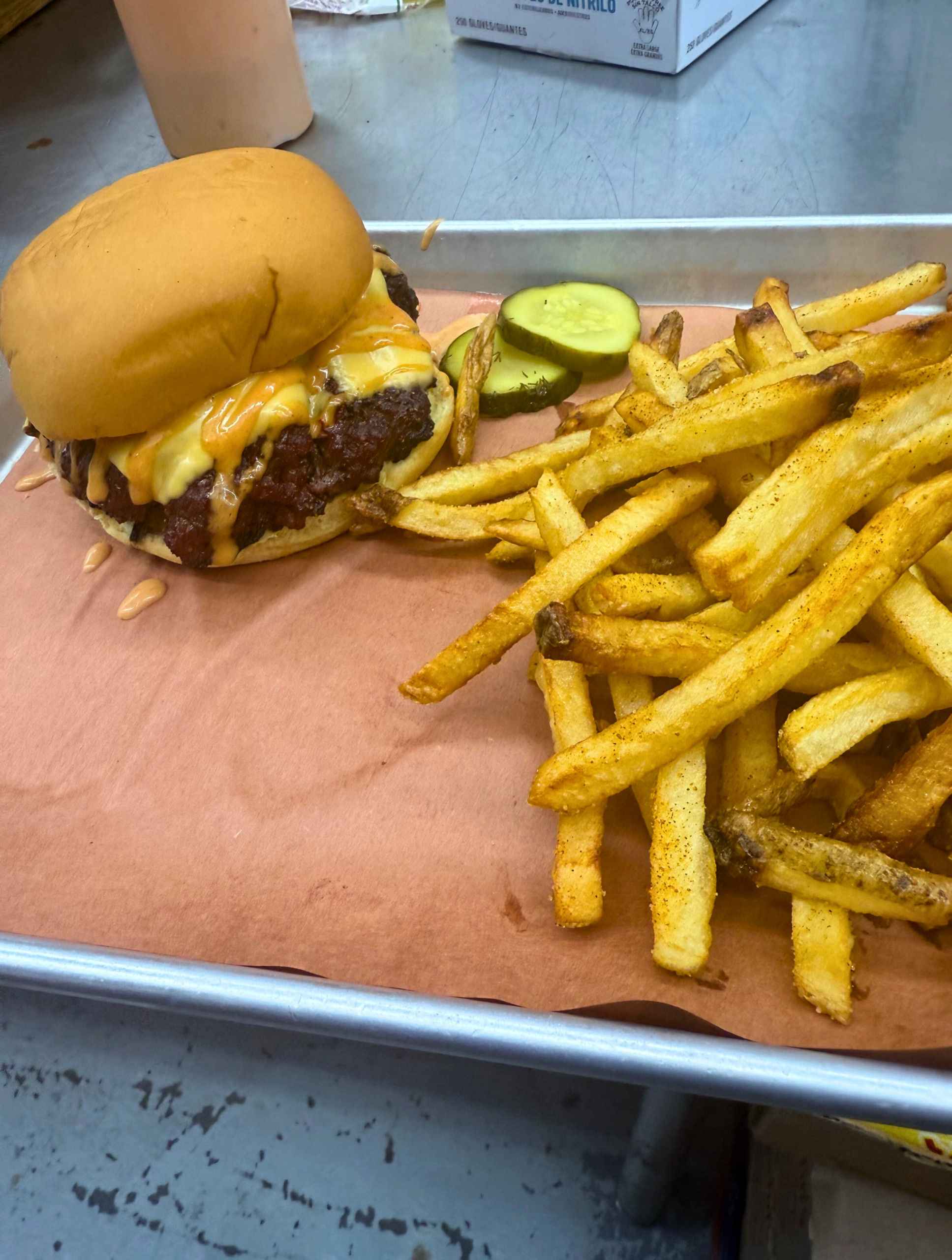 Ben Backdraft BBQ's Smokehouse Brisket Burger sits on a tin tray lined with parchment paper, accompanied by seasoned fries and sliced pickles.