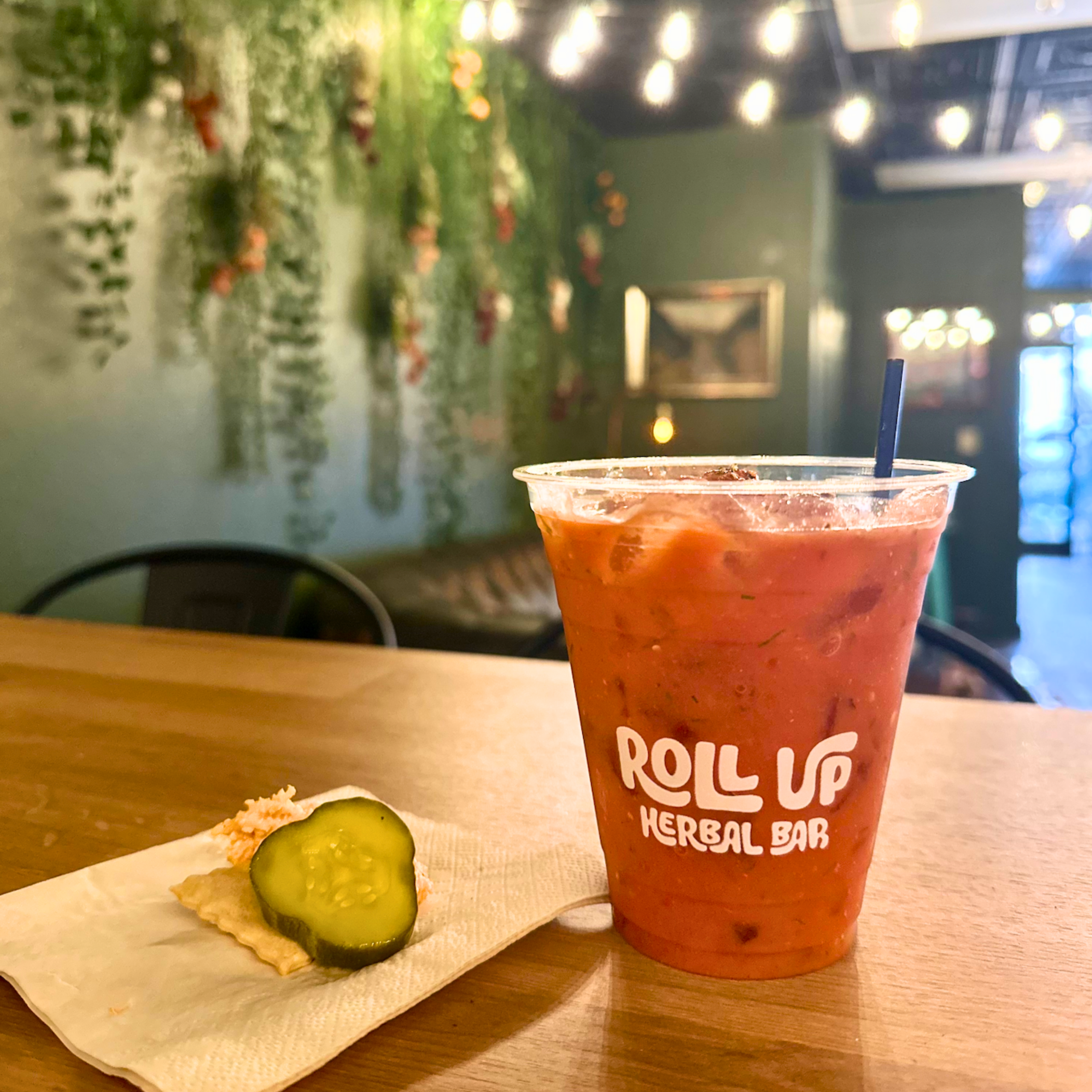 Roll Up Herbal Bar's "Something Burger" mocktail atop a wooden table, accompanied by a cracker bite topped with pickle and pimento cheese.